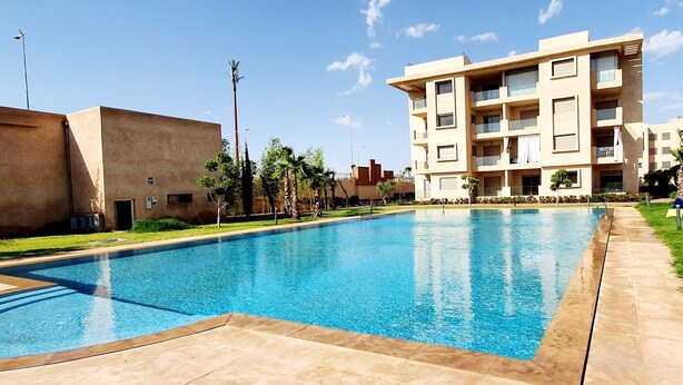 A rectangular swimming pool with clear blue water, surrounded by a tiled deck. In the background, there are multi-story buildings with balconies and a clear sky.