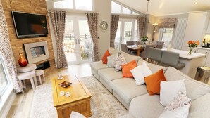 A spacious living room with a large sofa, a coffee table, and a TV mounted on the wall. There is a dining area with a table and chairs, and a kitchen area visible in the background. - 8 Waterside Wood (Windermere)
