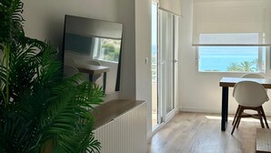 A modern living room with a TV, a dining table, chairs, a large potted plant, and a view of the sea through a window.