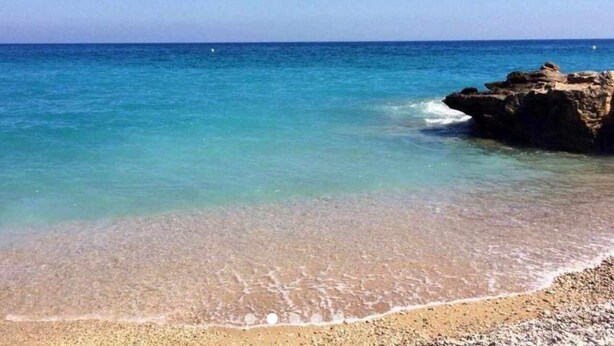 A beach with clear turquoise water, pebbly shore, and a rocky outcrop.