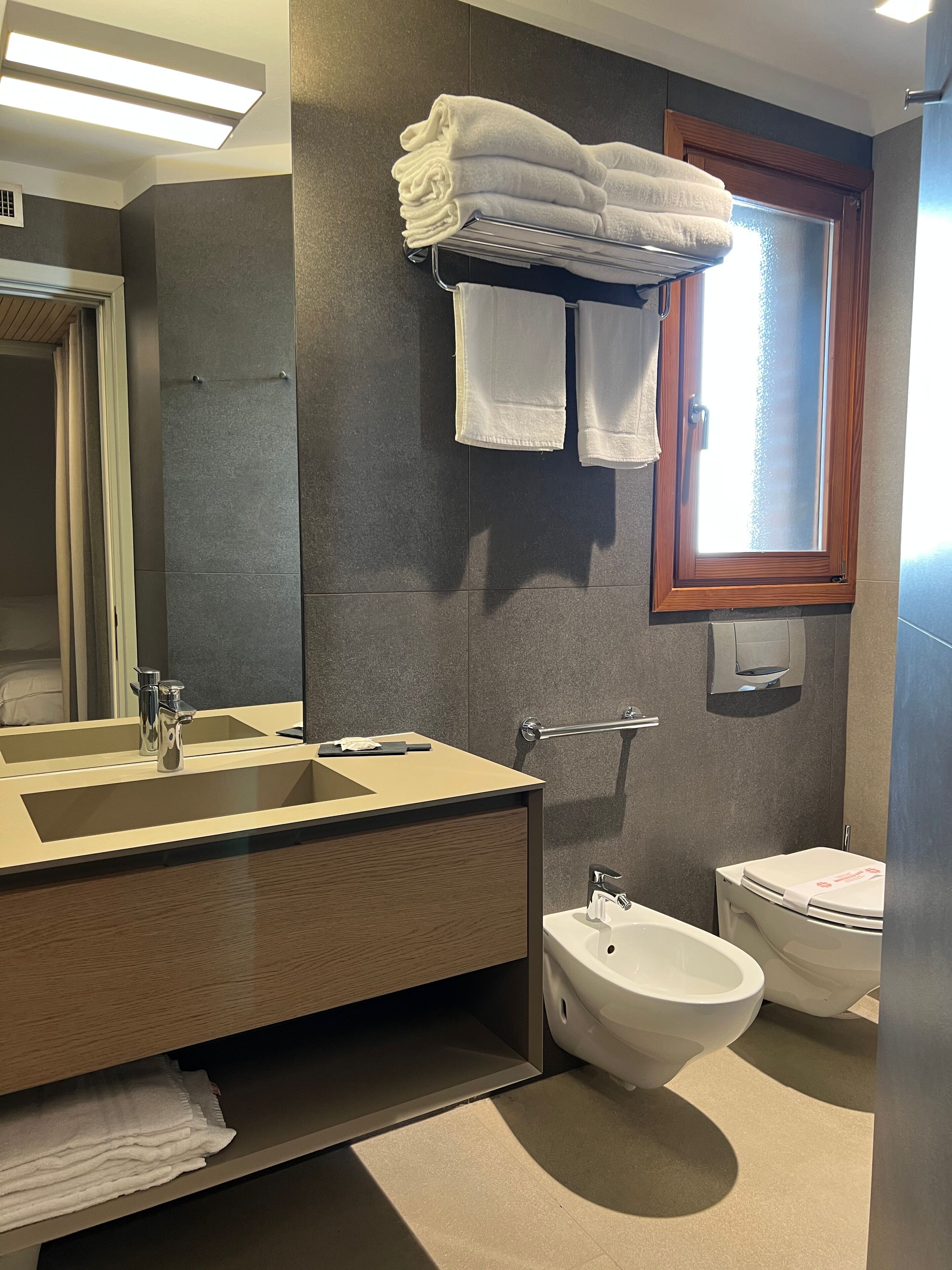 A modern bathroom with a large mirror, a sink, a bidet, and a toilet. There are towels stacked on a shelf and a window providing natural light.