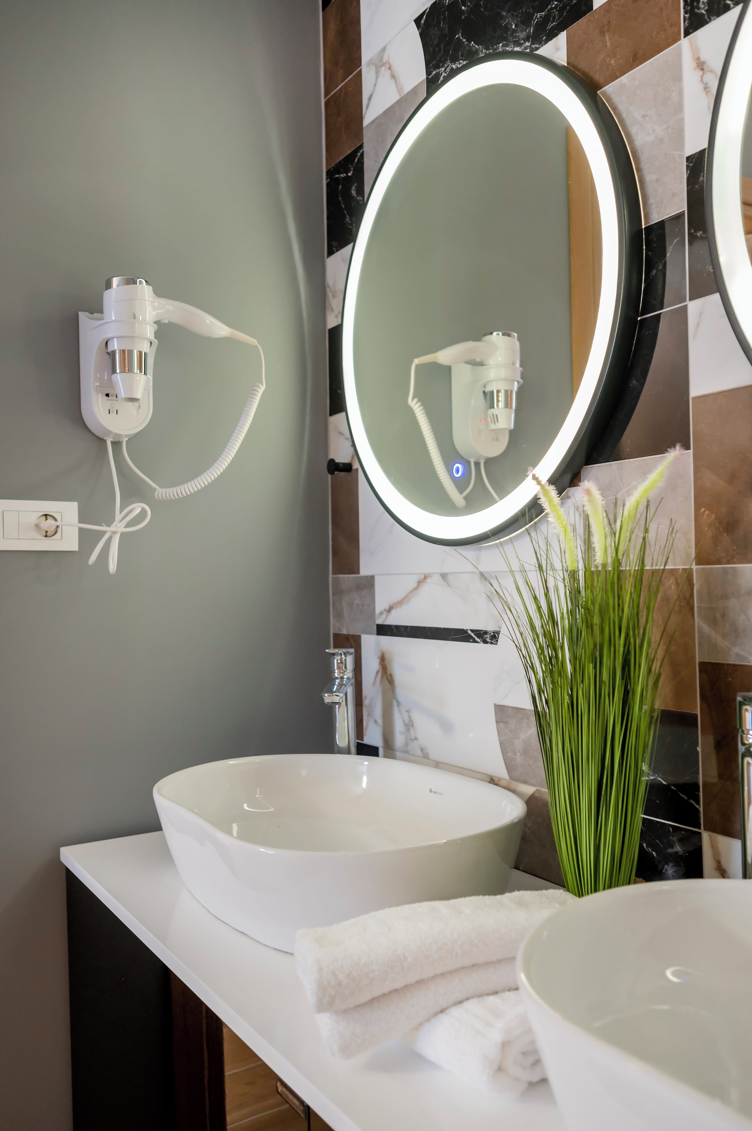 Een badkamer met twee witte wasbakken, een spiegel met verlichting, een haardroger en een groene plant.