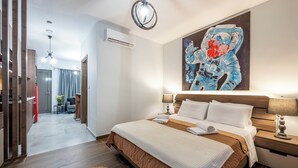 A modern hotel room with a large bed, a bedside table, a wall-mounted painting, and a view into the adjacent room.