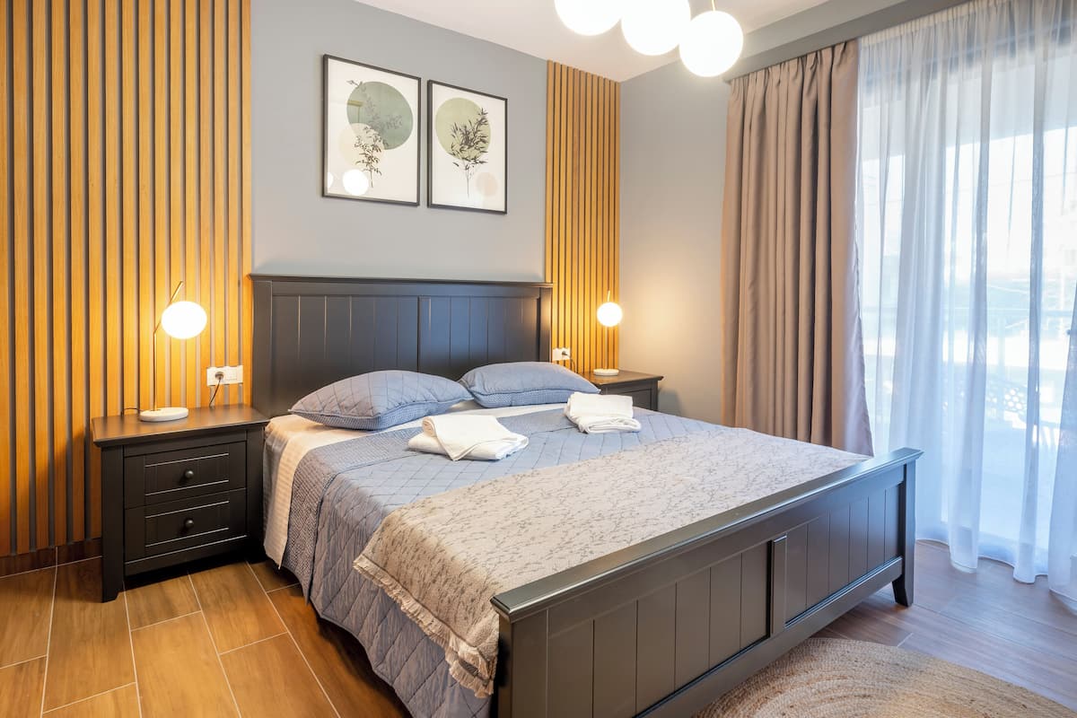 Een netjes opgemaakt bed met een blauwe quilt en witte handdoeken, een donker hoofdbord en twee bedlampen.