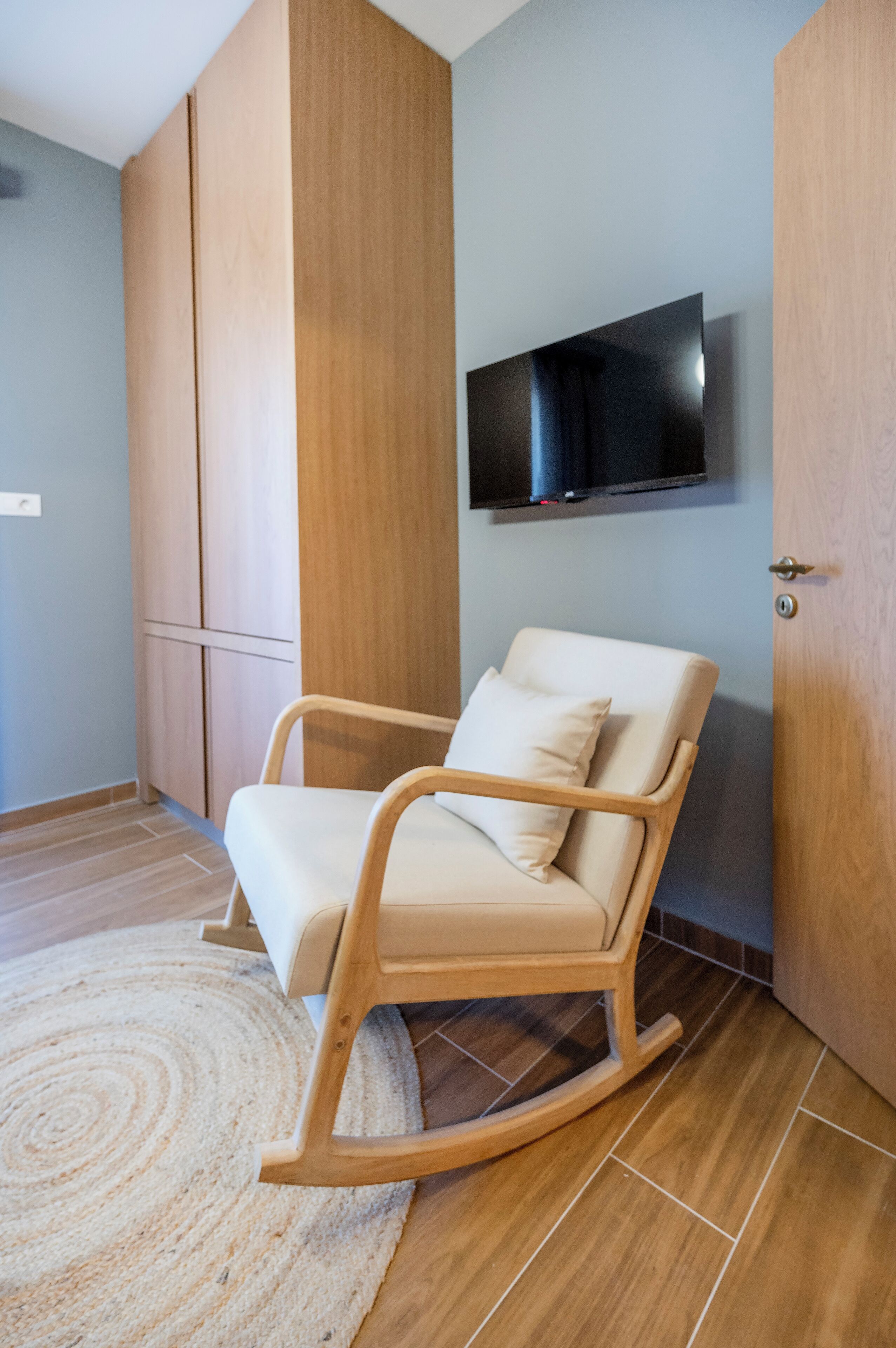 A room with a wooden rocking chair, a flat-screen TV mounted on the wall, and a wooden wardrobe.
