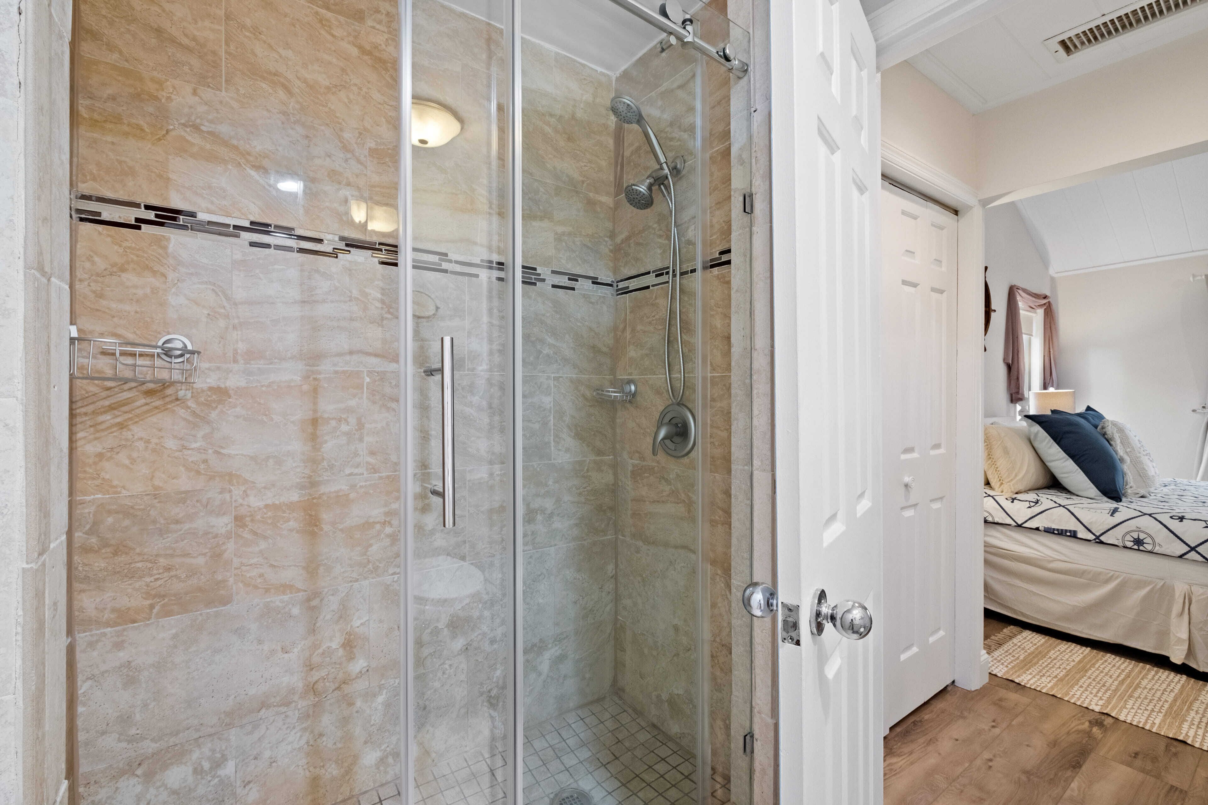 A modern bathroom with a glass-enclosed shower, a white door, and a bed with patterned bedding in the adjacent room.