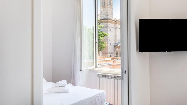 A hotel room with a bed, a window with a view of a tower, and a flat-screen TV mounted on the wall. - Corso View apartment (Rome)