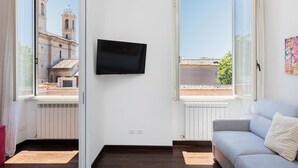 A room with a large window, a flat-screen TV mounted on the wall, and a grey sofa. - Corso View apartment (Rome)