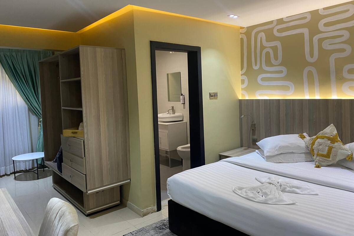 A modern hotel room with a large bed, a wardrobe, a bathroom, and a small seating area.