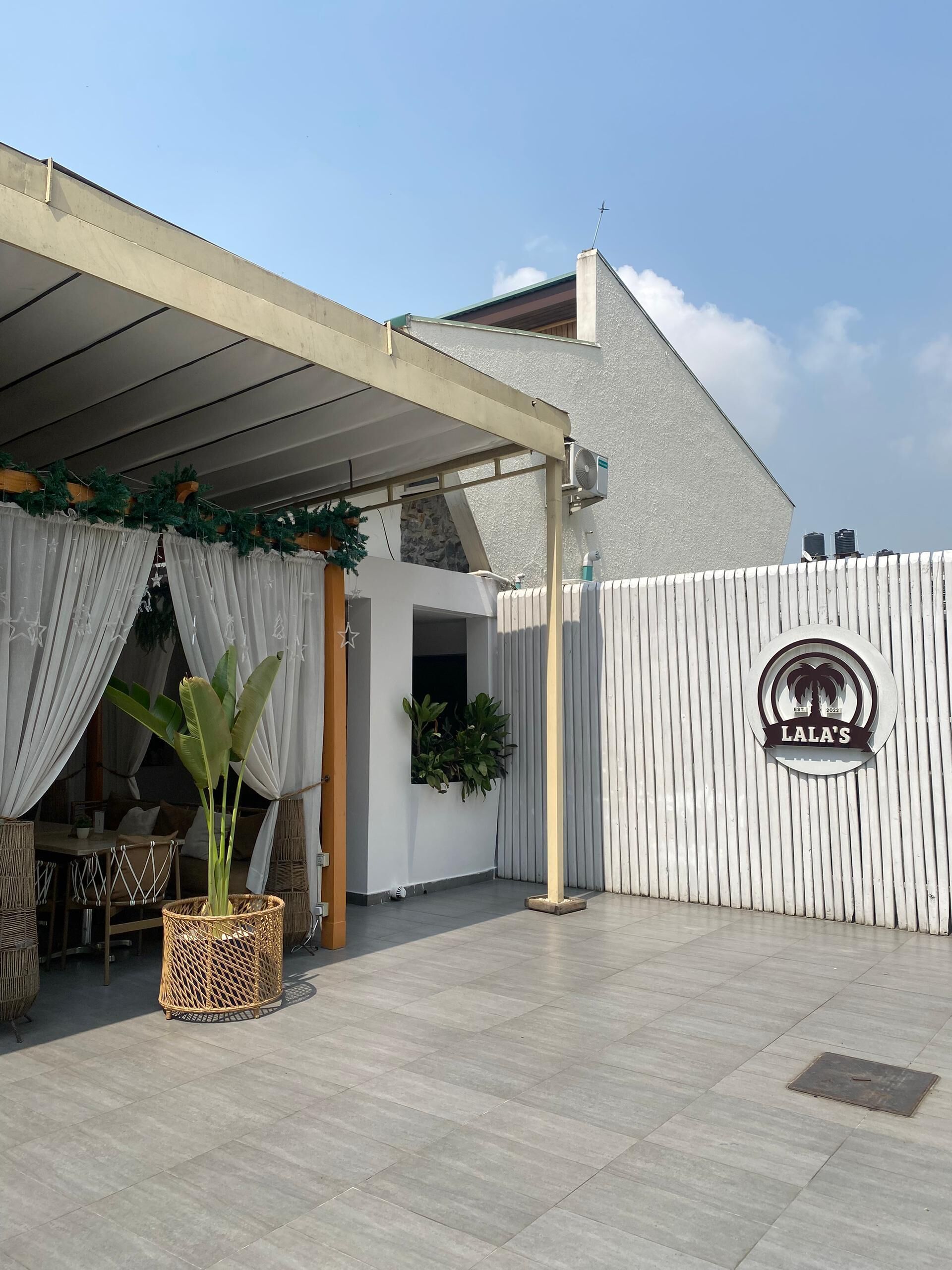 A covered outdoor seating area with a white fence, a sign reading "Lala's", and potted plants.