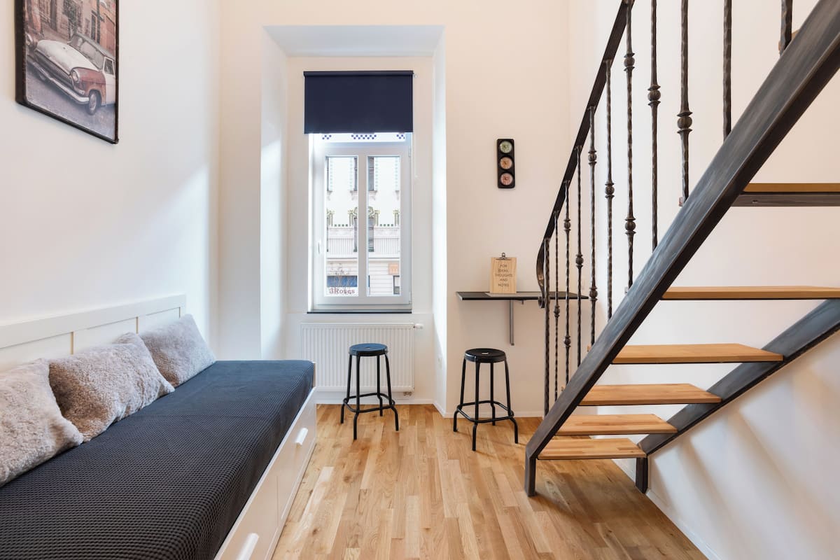 Un apartament modern amb un terra de fusta, un sofà negre, una escala de fusta i una finestra amb vista a edificis.