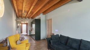 Ein Wohnzimmer mit Holzbalkendecke, einem gelben Sessel, einem dunklen Sofa und einer Küche im Hintergrund.