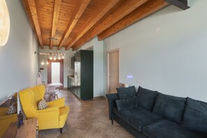 Una sala de estar con techo de madera, un sillón amarillo, un sofá oscuro y una cocina al fondo.