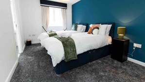 Una camera da letto moderna con una parete decorata in blu scuro, un letto grande con molti cuscini, un comodino con una lampada e una finestra con tende.