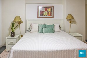A neatly made bed with a striped bedspread, two bedside tables with lamps, a framed artwork above the bed, and a potted plant on the left side. - Las Palmas (Mazatlán)