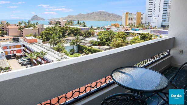 Un balcon avec une table et des chaises, d’où l’on peut admirer un panorama urbain avec des immeubles et une montagne à l’horizon.