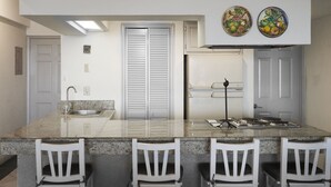 Une cuisine dotée d’un plan de travail en granit, de chaises blanches, d’une porte à deux vantaux et d’un four encastré.