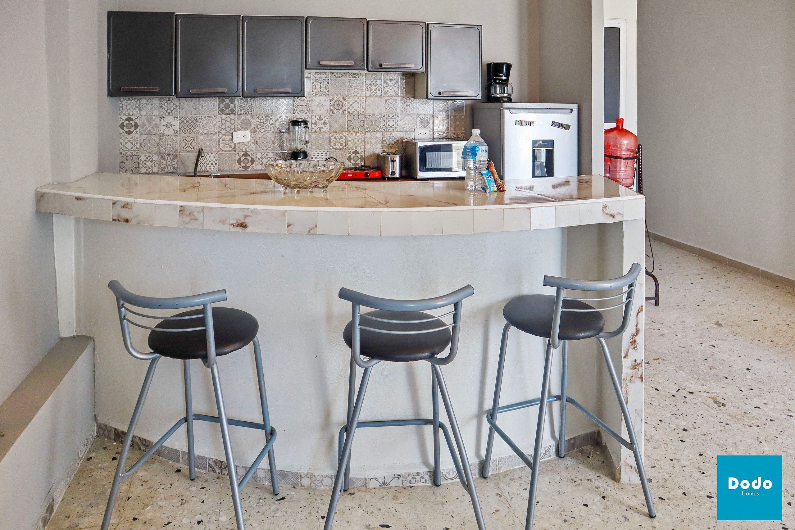 Una cocina con encimera de mármol, máquina de café, dispensador de agua y tres taburetes de bar.