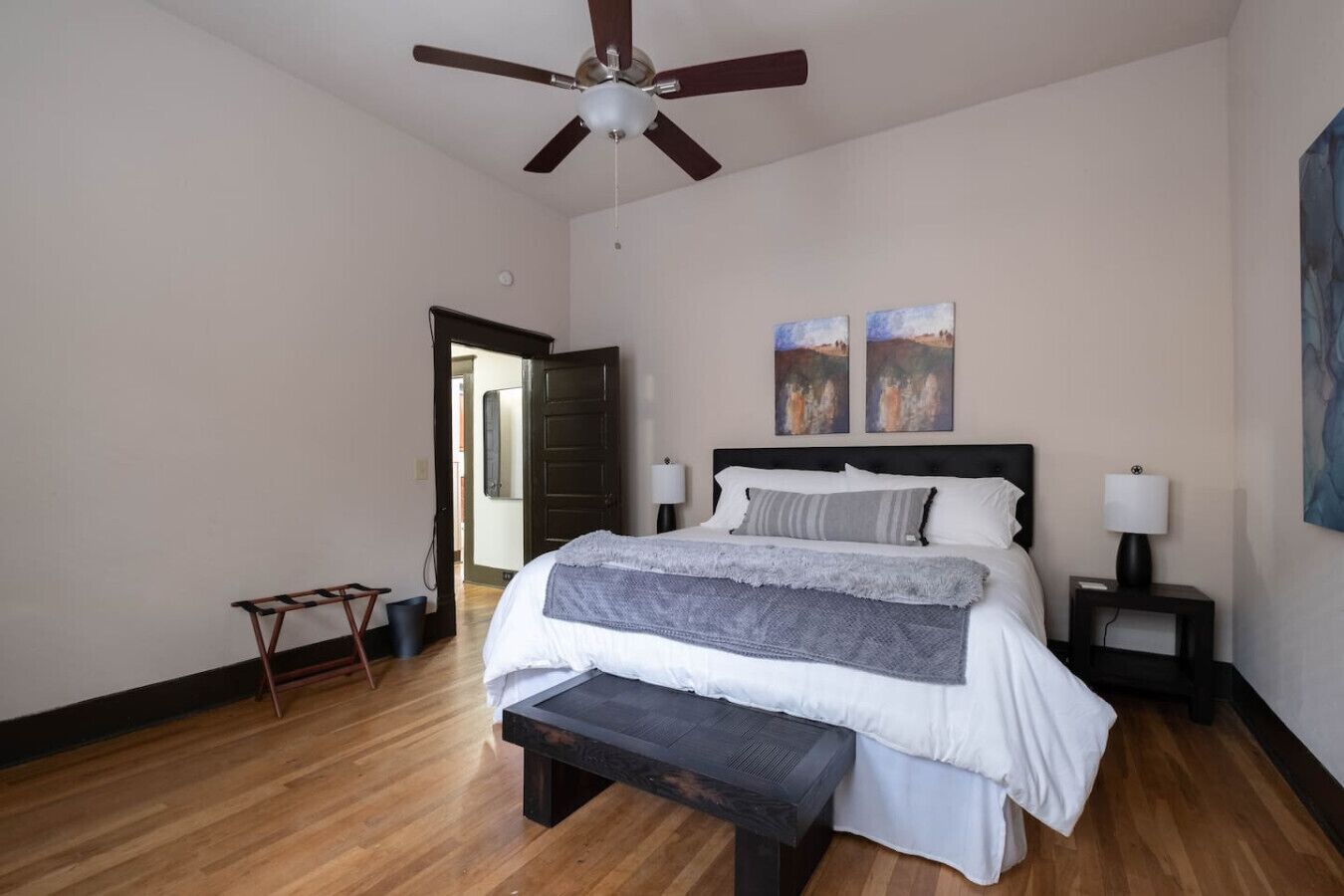 A bedroom with a bed, a ceiling fan, two bedside lamps, a bench, and two paintings on the wall.