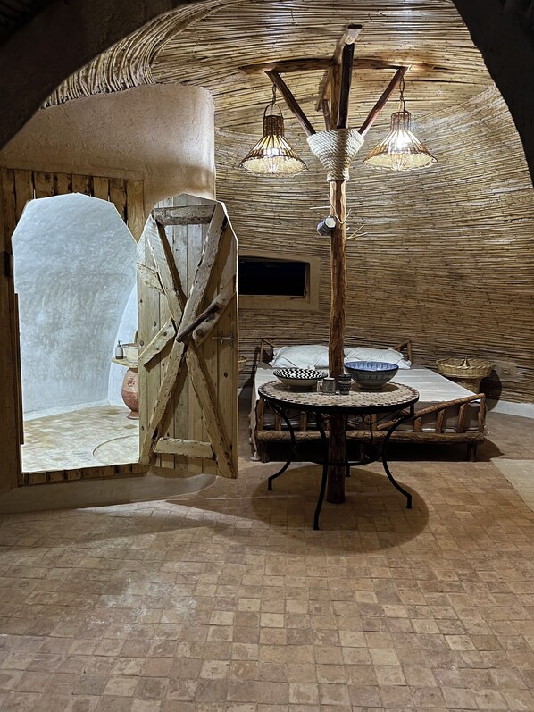 A rustic interior with a round table, wicker chairs, a woven ceiling, and a wooden door.