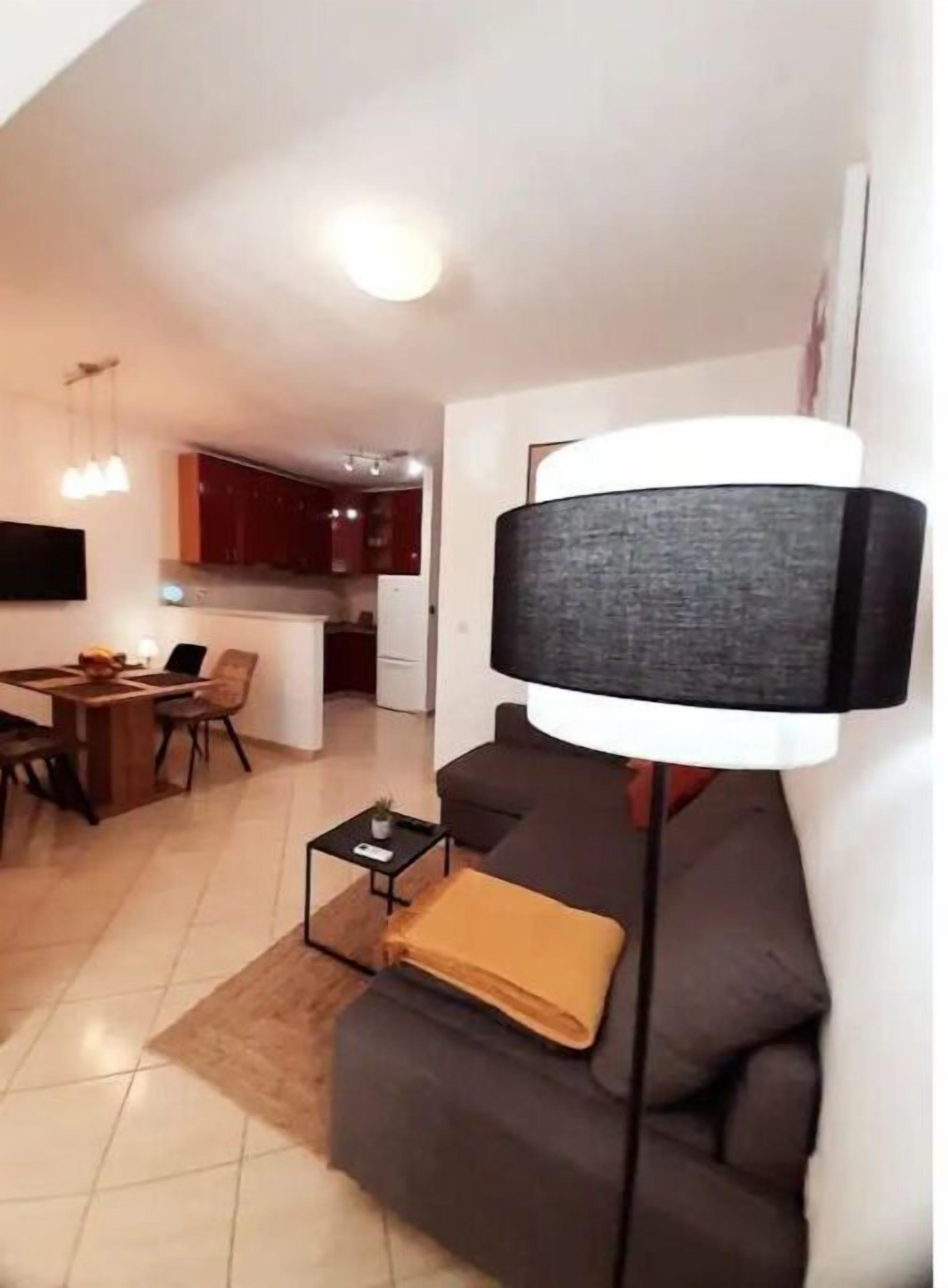 Un salon moderne avec un canapé foncé, une table d’appoint et une lampe sur pied. On y trouve également un coin repas avec une table et des chaises, et une cuisine à l’arrière-plan.