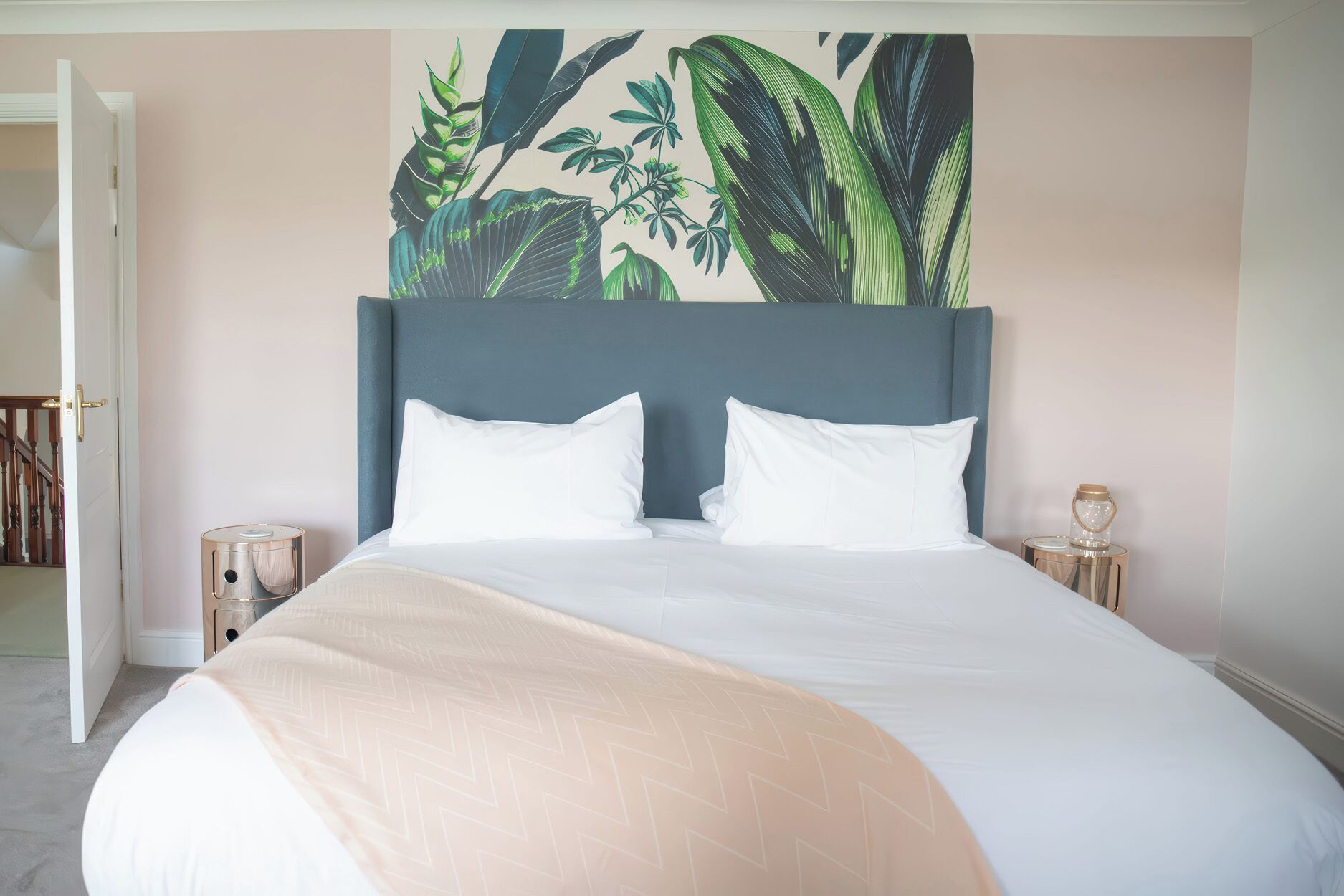A neatly made bed with white linens and a beige throw blanket, a blue headboard, and a wall with a tropical plant mural.