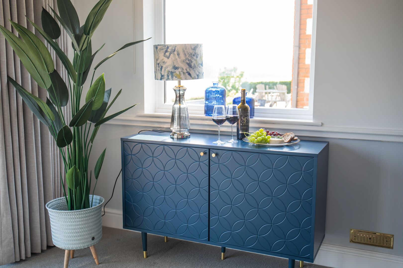 Een blauwe kast met een geometrisch patroon, een potplant, een lamp en een bord druiven en kaas bij een raam.