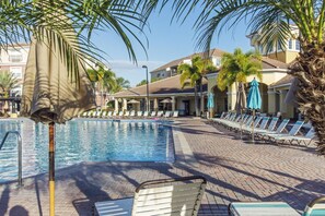 Un área de piscina con sillas de descanso y sombrillas, rodeada de palmeras y un edificio con tejado de baldosas.