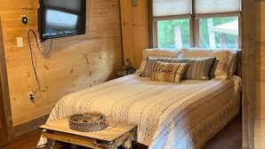 Une pièce chaleureuse, aménagée dans une cabane en bois, avec un lit, un téléviseur fixé au mur et une fenêtre équipée de stores.