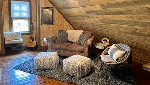 Une chambre à coucher confortable, aménagée en mezzanine, avec un plafond en pente, des murs en bois, un canapé en cuir marron, deux poufs en osier, une chaise en rotin et une guitare.