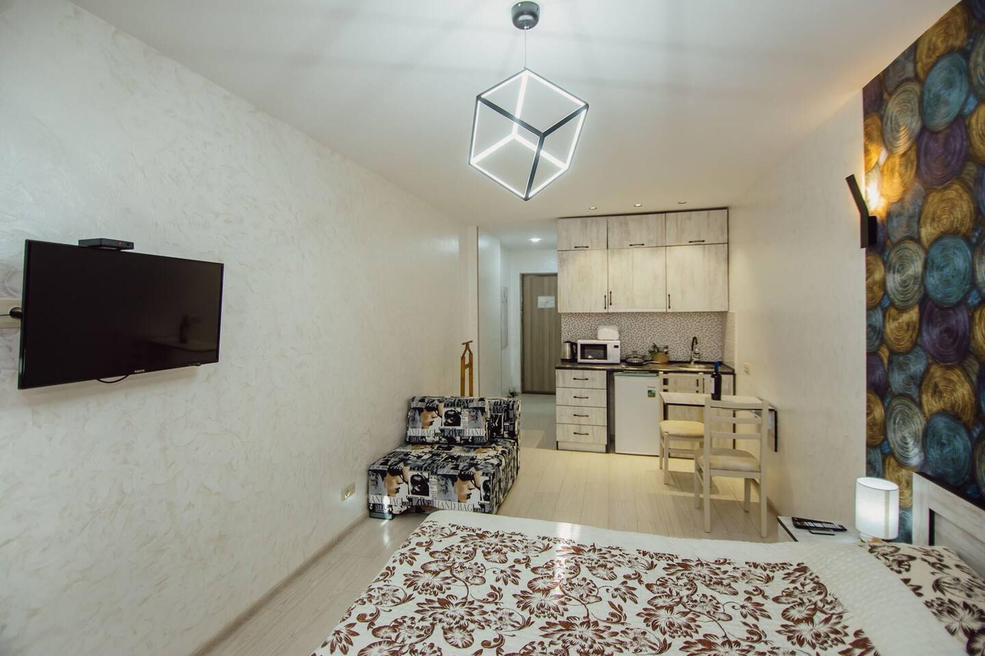 A bedroom with a bed, a TV, a unique geometric light fixture, a kitchenette, and a colorful tapestry.