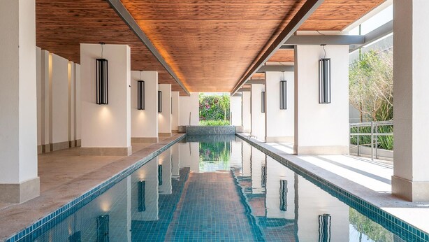 A long indoor swimming pool with a wooden ceiling and tiled walls, surrounded by columns and a view of greenery. - Is Moema (São Paulo)