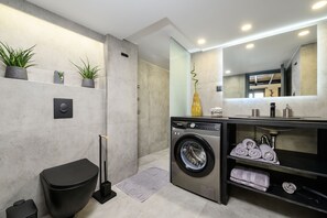 A modern bathroom with a grey tiled wall, a black toilet, a washing machine, and a shelf with towels. - Loft 1 (Kos)