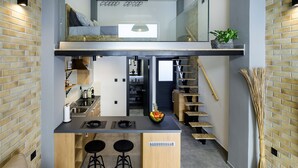 A modern kitchen with a central island, bar stools, and a staircase leading to a loft bedroom. - Loft 1 (Kos)