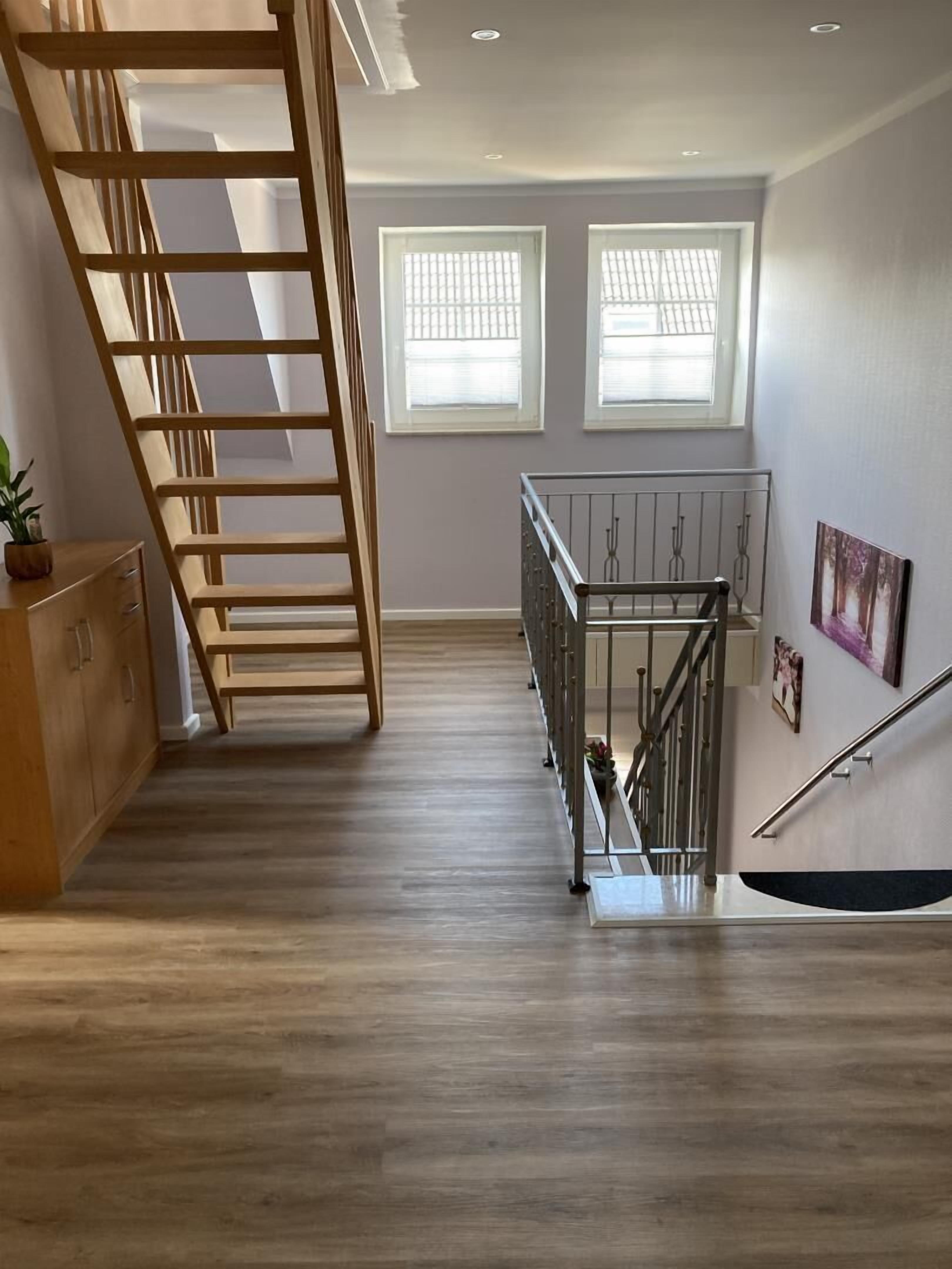 Een ruime kamer met een houten vloer, een houten trap, een metalen leuning en twee ramen met zonwering.