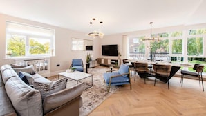 Un vaste salon avec un grand canapé, un coin repas comprenant une table et des chaises, un téléviseur à écran plat fixé au mur et de grandes fenêtres donnant sur la verdure.