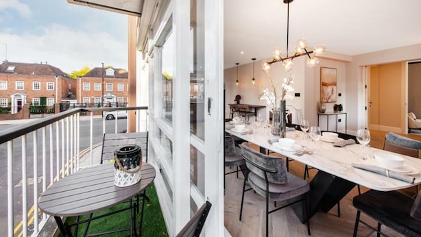 Un appartement moderne doté d’un balcon, d’un coin repas avec une longue table dressée pour quatre personnes, et offrant une vue sur un immeuble en briques situé de l’autre côté de la rue.