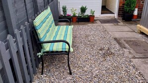 Un banc avec un coussin à rayures, placé dans un espace recouvert de gravier, une clôture en bois et une porte blanche avec des fenêtres.