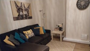 Un salon avec un canapé d’angle, un tapis à motifs, une horloge murale et un tableau représentant des ânes.