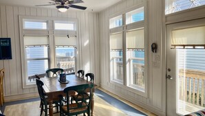 Ruokailutilassa on puinen pöytä ja tuolit, suuret ikkunat, joissa on sälekaihtimet, sekä kattotuuletin.