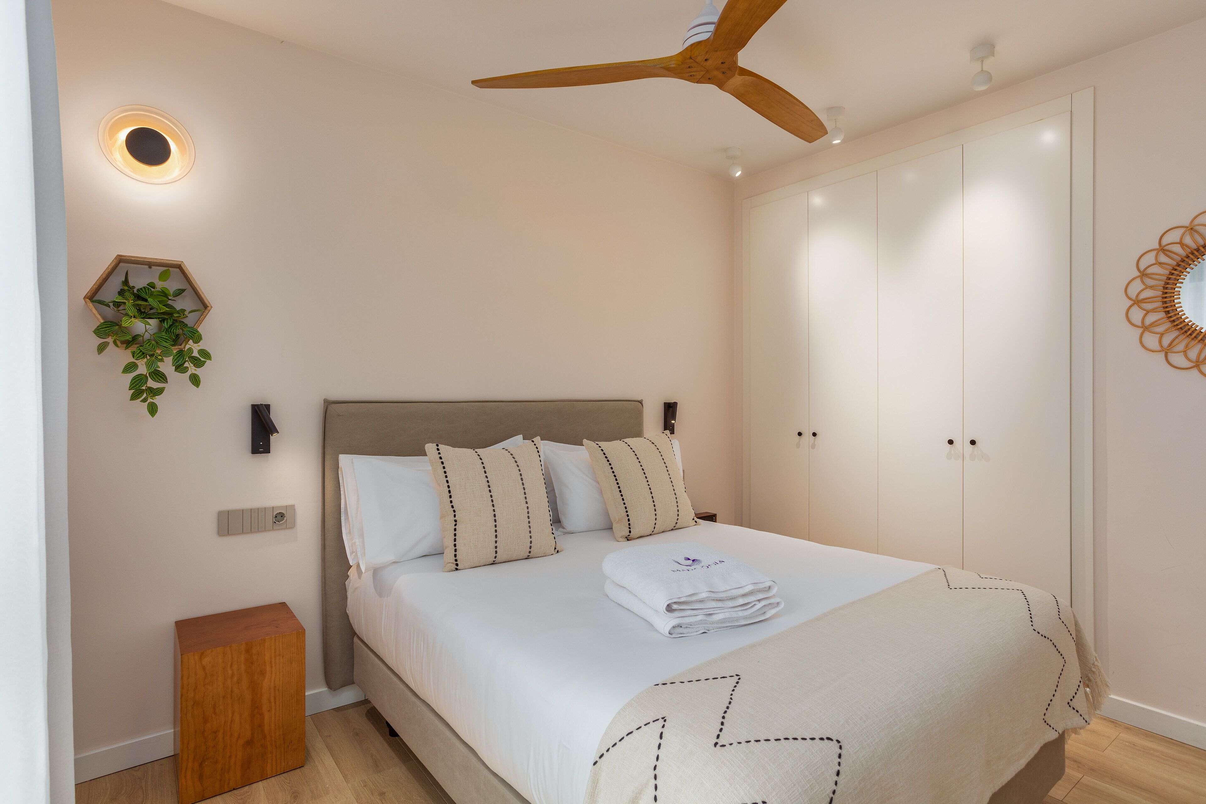 Un dormitorio con una cama, un ventilador de techo, una lámpara fijada a la pared, una mesita de madera y una planta en maceta.