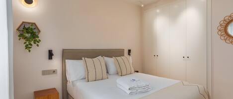 Un dormitorio con una cama, un ventilador de techo, una lámpara fijada a la pared, una mesita de madera y una planta en maceta.