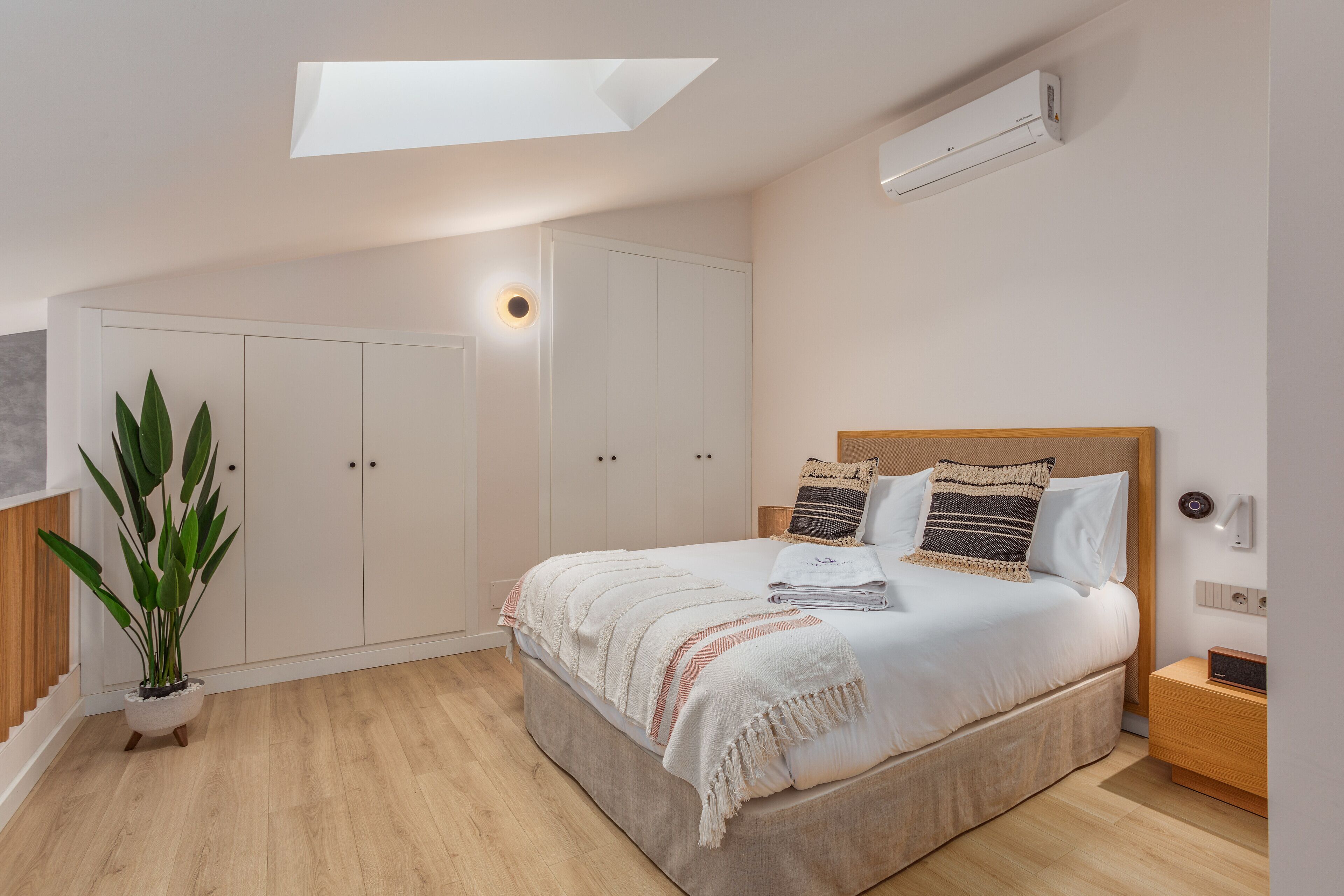 Un dormitorio con tragaluz, una cama con almohadas, una mesita de noche y un armario, además de una planta en maceta.
