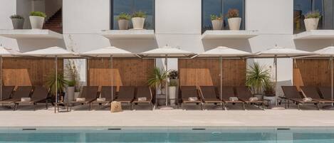 Zona junto a la piscina con sillones de descanso y sombrillas, una terraza de madera y plantas en macetas en la fachada del edificio.