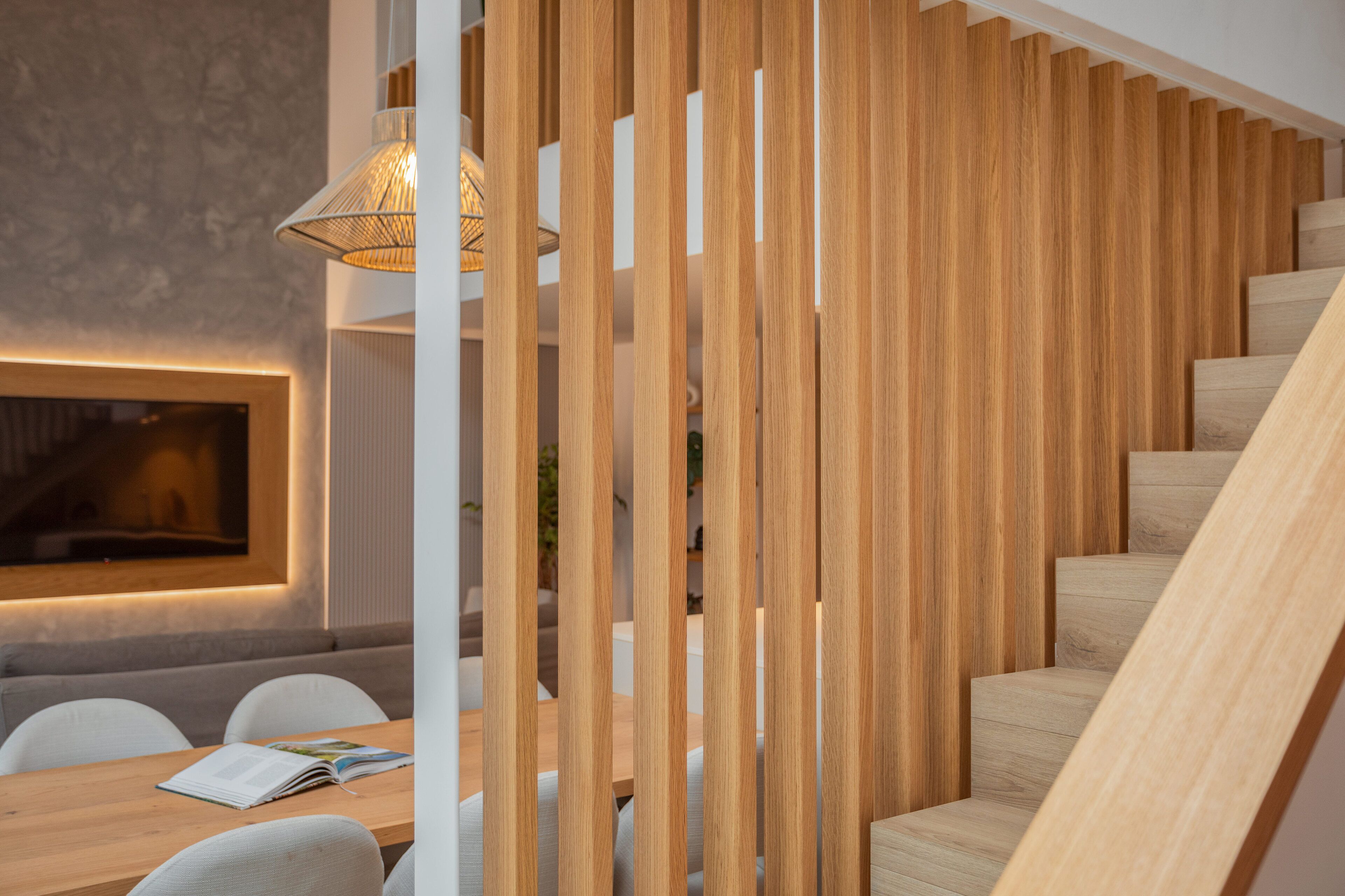 Un interior moderno con una escalera de madera, una zona de comedor con una mesa y sillas, y un aplique de pared.