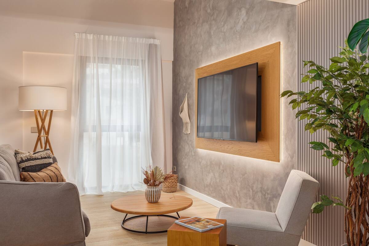 Ein modernes Wohnzimmer mit einem runden Couchtisch aus Holz, einem grauen Sofa, einem weißen Sessel und einem großen Flachbildfernseher, der an der Wand befestigt ist.