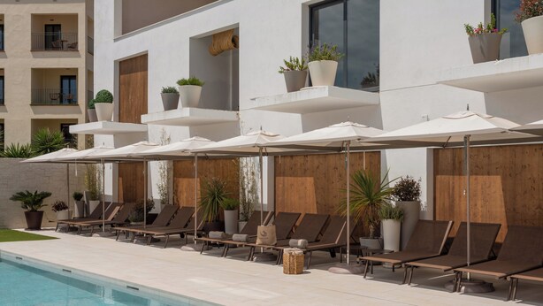 Un área junto a la piscina con sillones de descanso y sombrillas, plantas en macetas y fachada de un edificio moderno.