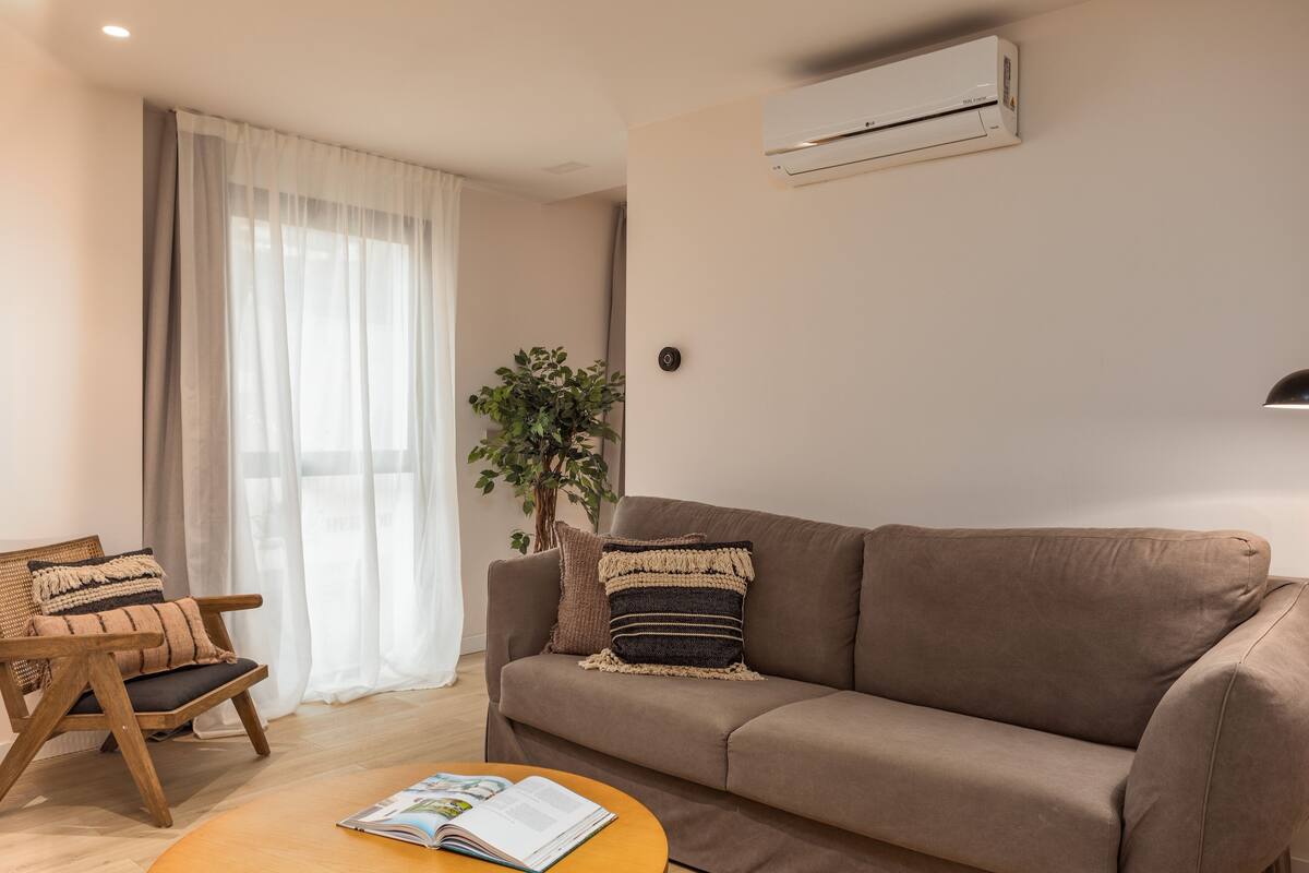 Ein Wohnzimmer mit braunem Sofa, einem Holzstuhl, einem runden Couchtisch mit einer Zeitschrift, einer Topfpflanze und einem großen Fenster mit transparenten Vorhängen.