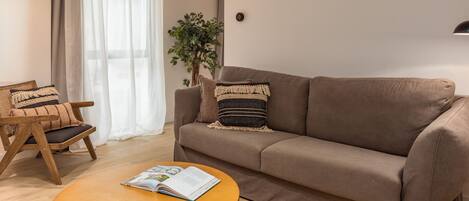A living room with a brown sofa, a wooden chair, a round coffee table with a magazine, a potted plant, and a large window with sheer curtains.