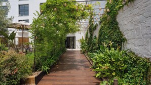Un sendero de madera bordeado por abundante vegetación y un edificio blanco con numerosas ventanas.